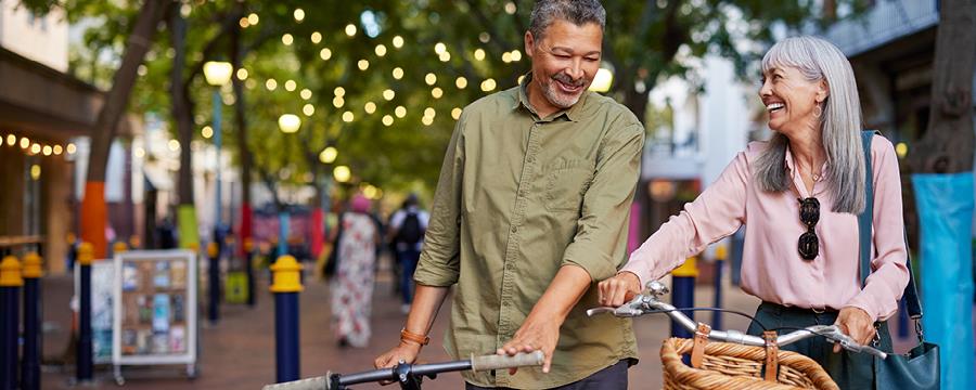 Retired couple walking their bikes on a city street.