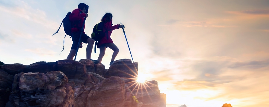 Two people stand on top of a mountain at sunset with packs and a walking stick
